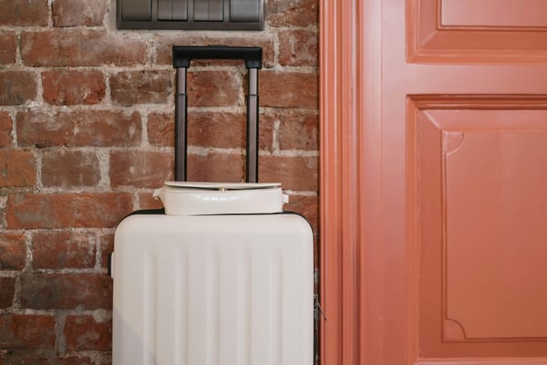 White rolling suitcase against red brick wall and coral-colored door with light switches