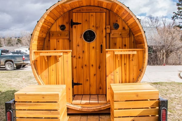 Wooden barrel-shaped glamping pod with rounded roof, central door, and wooden steps, parked on gravel with bare trees and pickup truck visible behind.