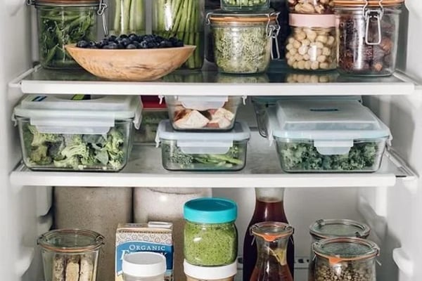 Organized refrigerator shelves with fresh vegetables, grains, legumes, and condiments in clear glass and plastic containers