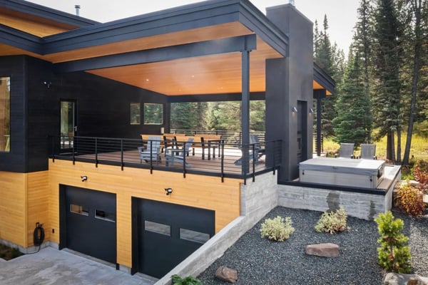 Modern mountain home with wood and dark exterior, two-car garage, upper deck, surrounded by pine forest and mountains