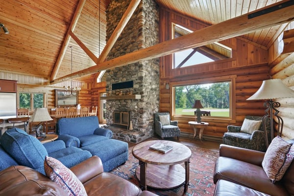 Rustic log cabin with front porch and bench, surrounded by evergreen trees and grassy lawn with forested hillside backdrop